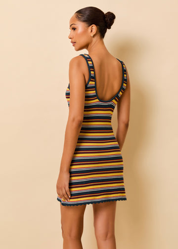 A woman modeling a sleeveless, open-back mini dress with horizontal navy, red, yellow, and white stripes, shown from a side-back angle against a solid beige studio background.