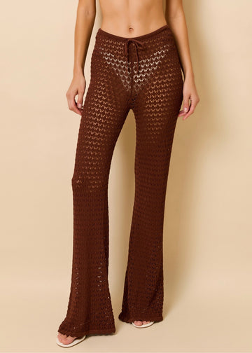A close up of a woman modeling a dark brown crochet wide-leg knit pants, set against a solid beige studio background.