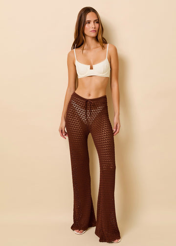 A woman modeling a cream-colored bikini and dark brown crochet wide-leg knit pants, set against a solid beige studio background.