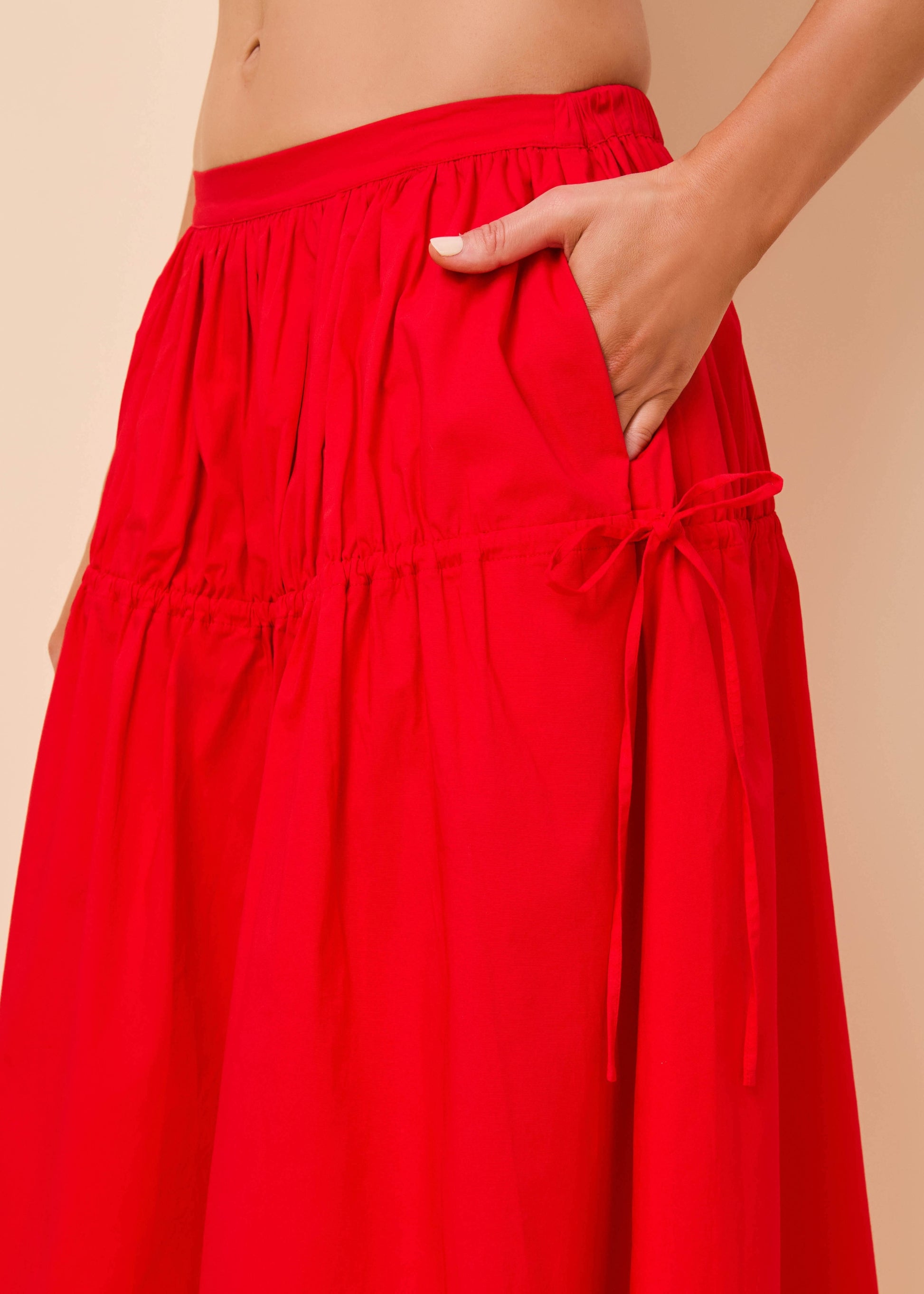 A close-up of a bright red tiered maxi skirt with side pockets and drawstring ties, shown on a model against a warm beige background.