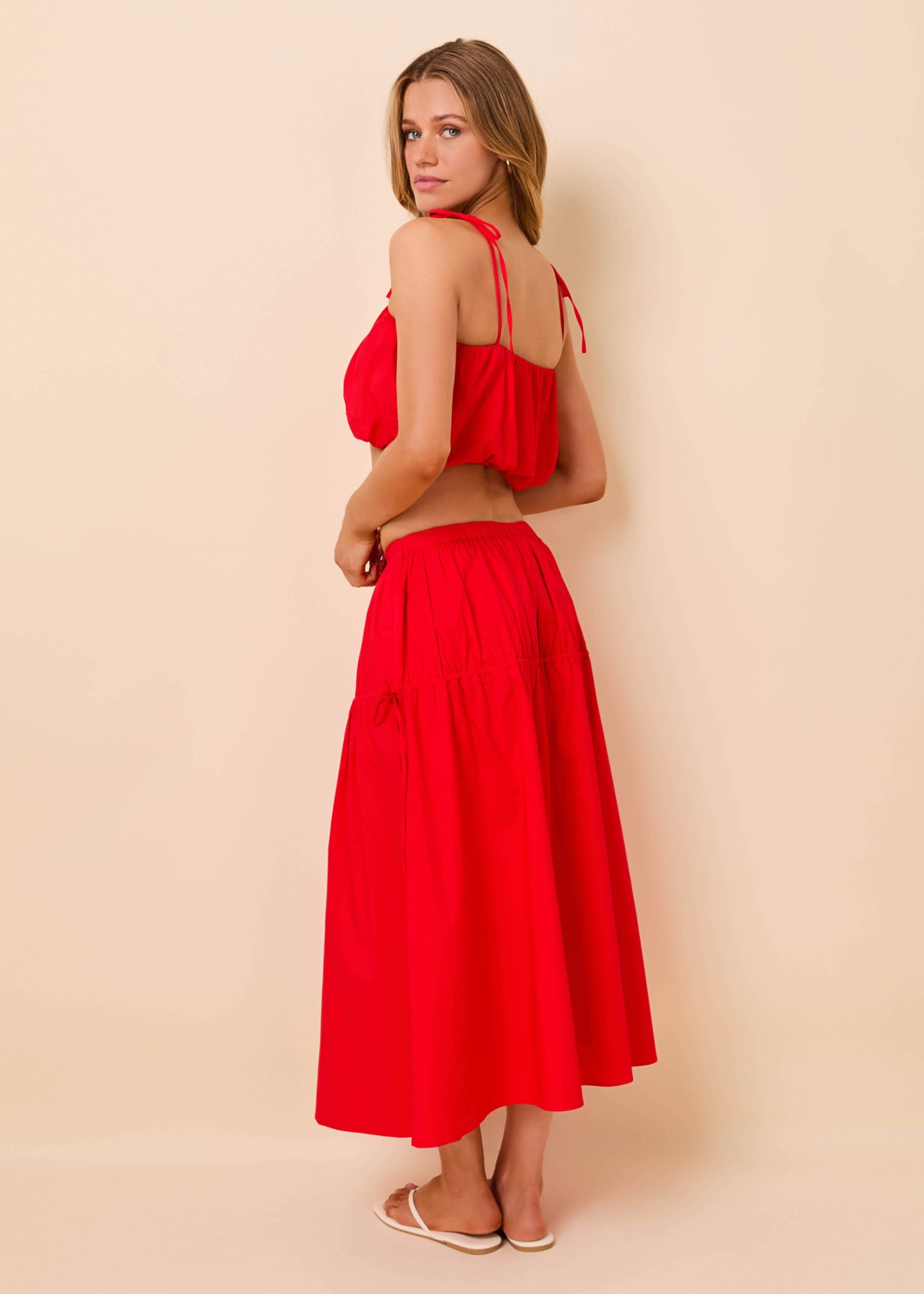 A woman modeling a bright red two-piece outfit consisting of a halter crop top and a matching high-waisted midi skirt, shown from a three-quarters back view against a solid beige studio background.