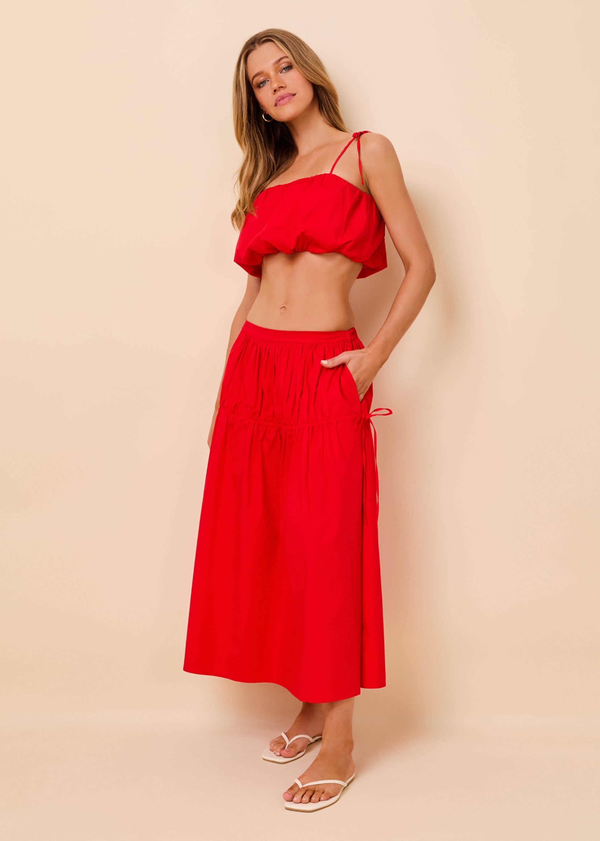 A woman modeling a bright red two-piece set consisting of a ruffled spaghetti-strap crop top and a matching high-waisted maxi skirt with side pockets, paired with white slide sandals, set against a solid beige studio background.