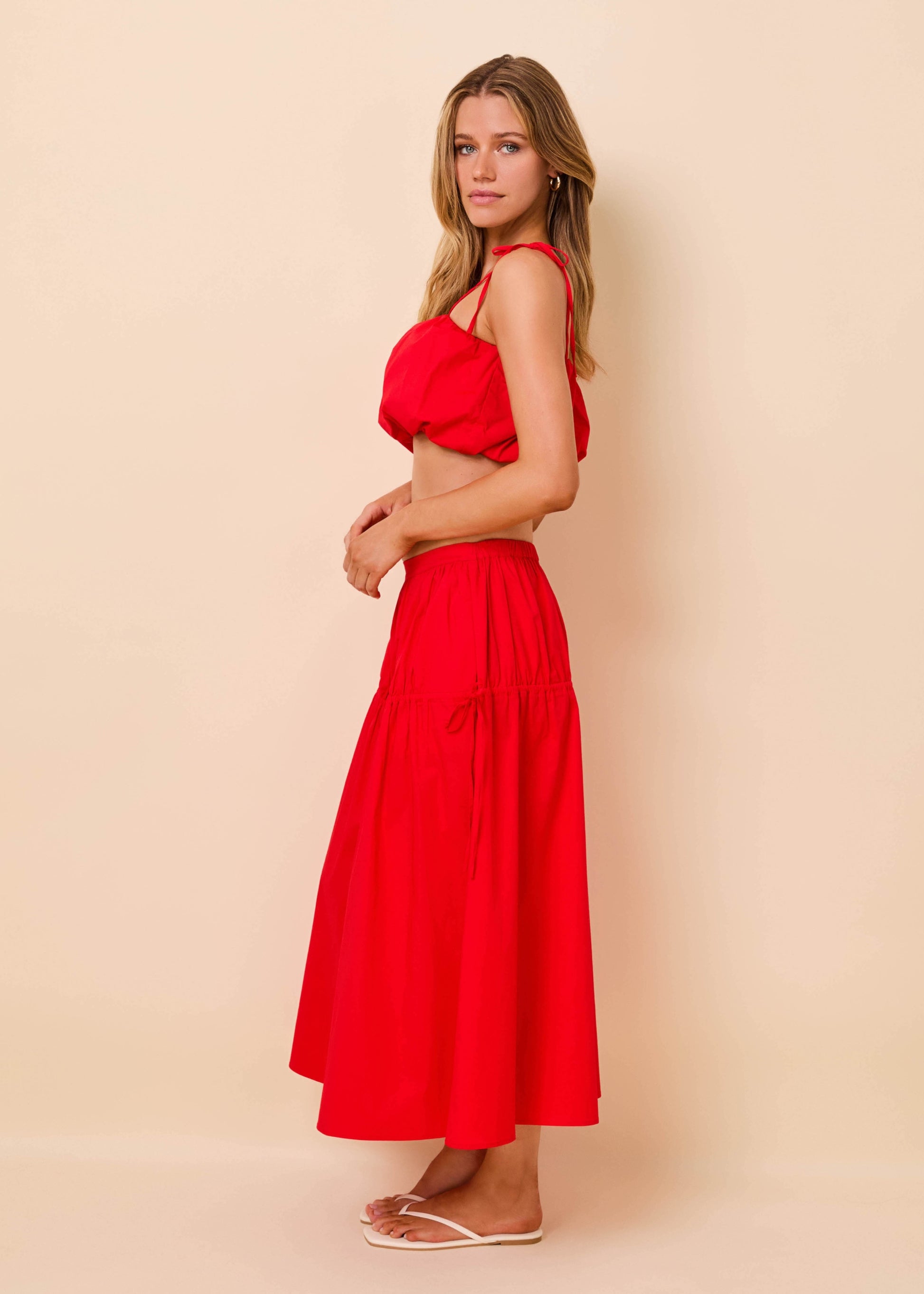A woman modeling a bright red two-piece set consisting of a sleeveless crop top with ruffled shoulders and a matching high-waisted maxi skirt, posed against a solid light beige background.