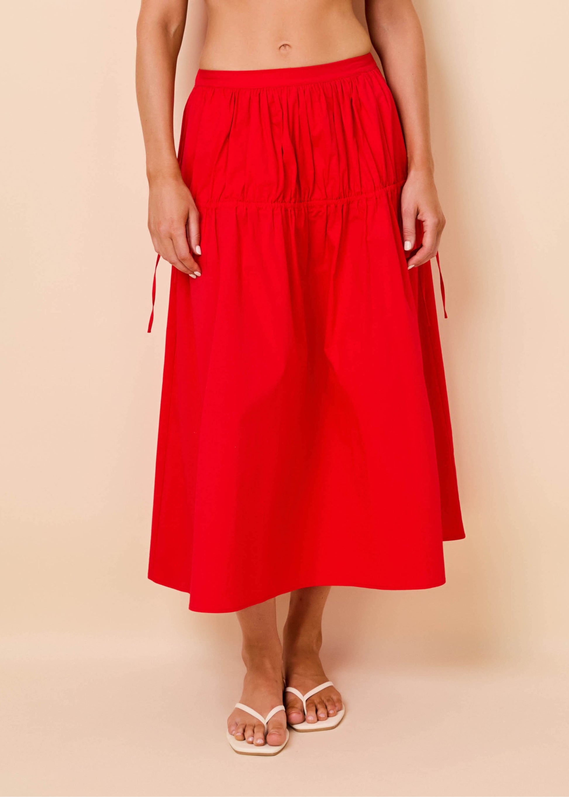 A bright red midi skirt with a gathered waist and side drawstrings, shown on a model wearing white slide sandals against a solid beige background.