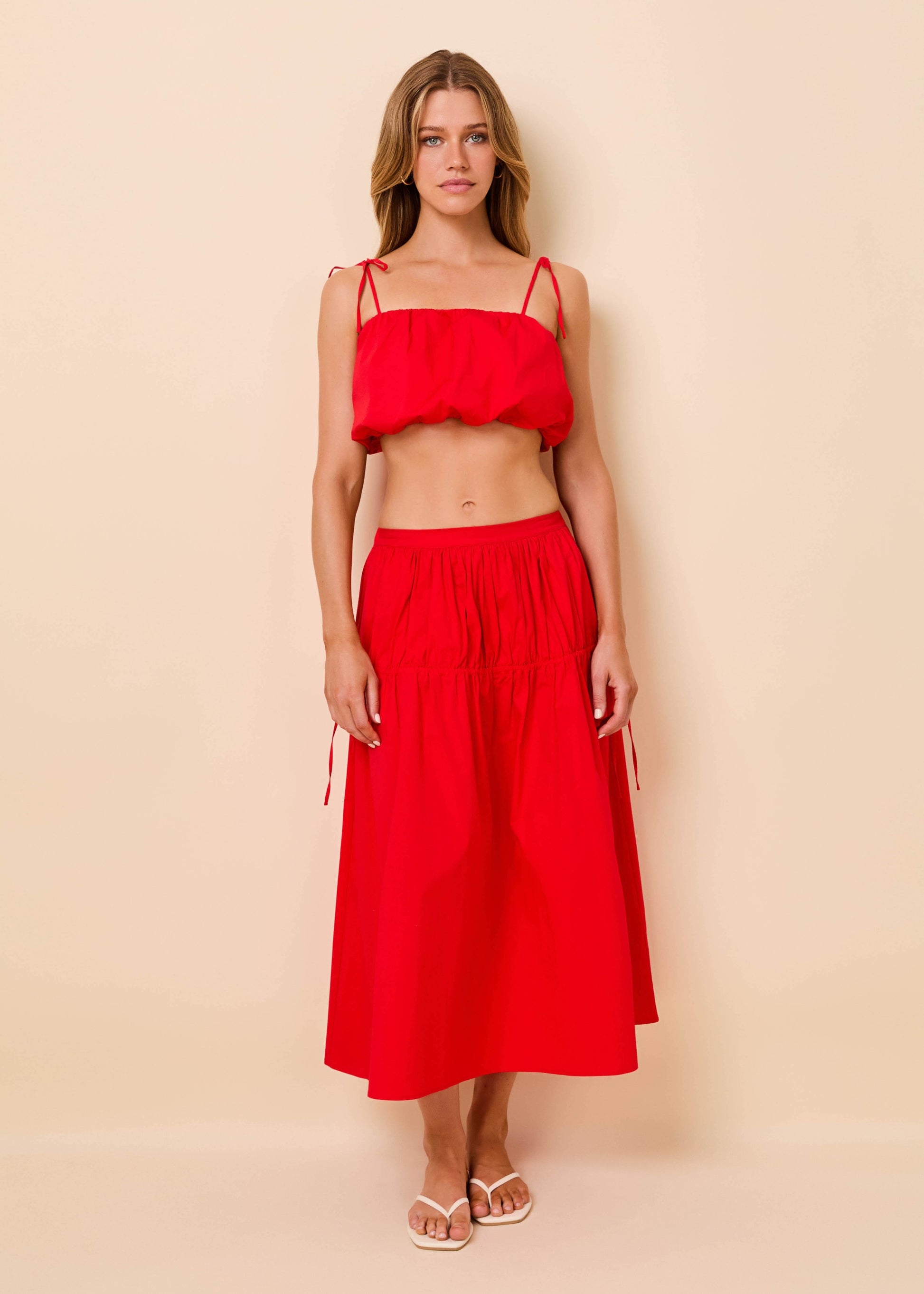 A woman modeling a bright red two-piece set consisting of a ruffled spaghetti-strap crop top and a matching high-waisted midi skirt with tie details at the hips, set against a solid light beige background.