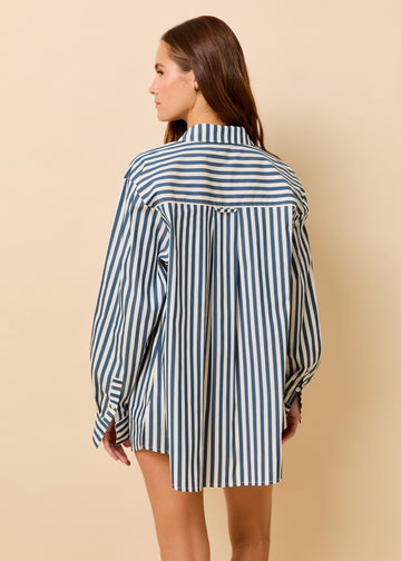 A woman shown from the back wearing an oversized long-sleeve button-up shirt dress with vertical navy and white stripes, set against a solid beige studio background.