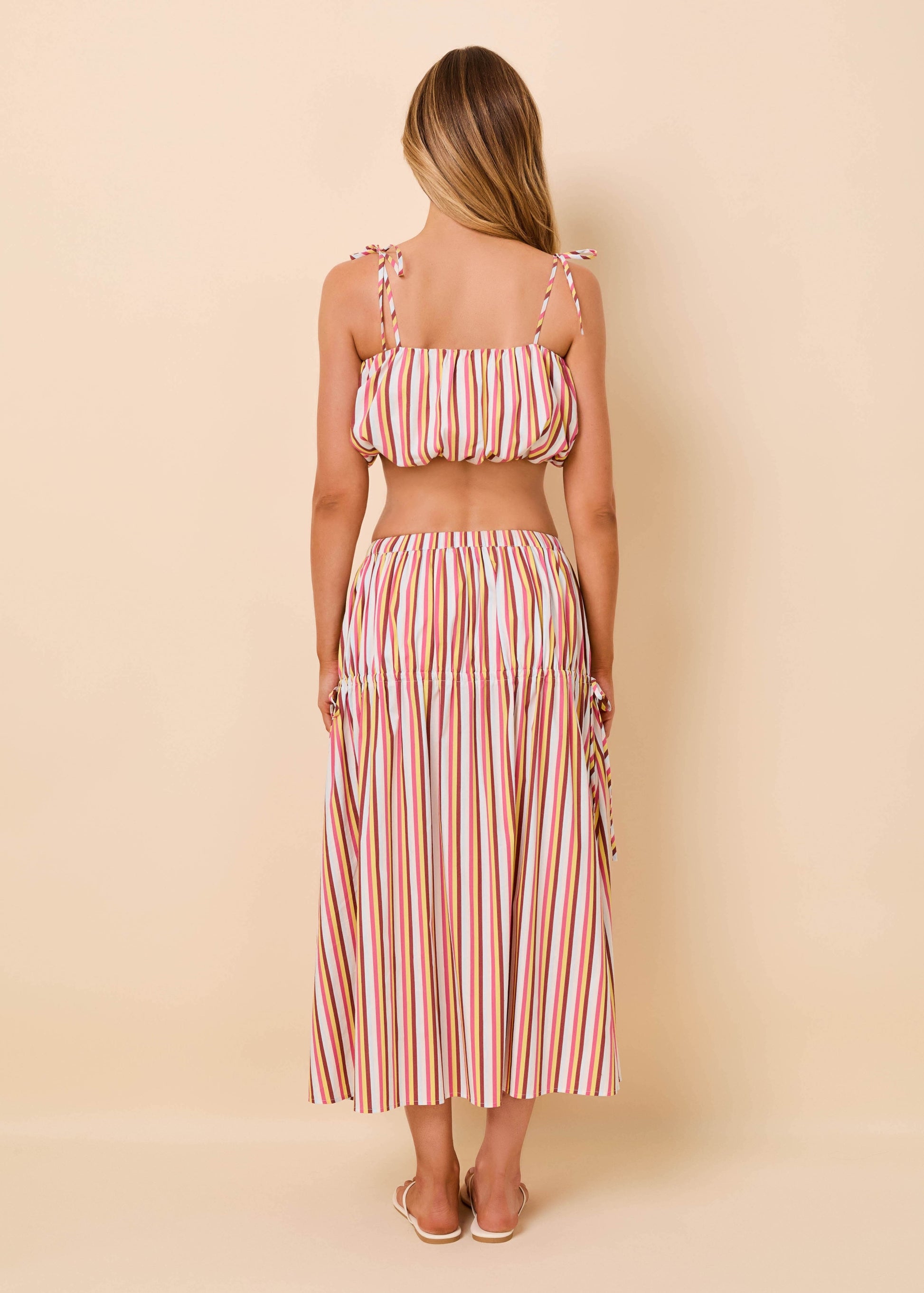 A woman shown from the back wearing a matching two-piece set with a cropped tie-strap top and a long pleated skirt, both in a vertical pink, orange, and white stripe pattern, set against a solid beige background.