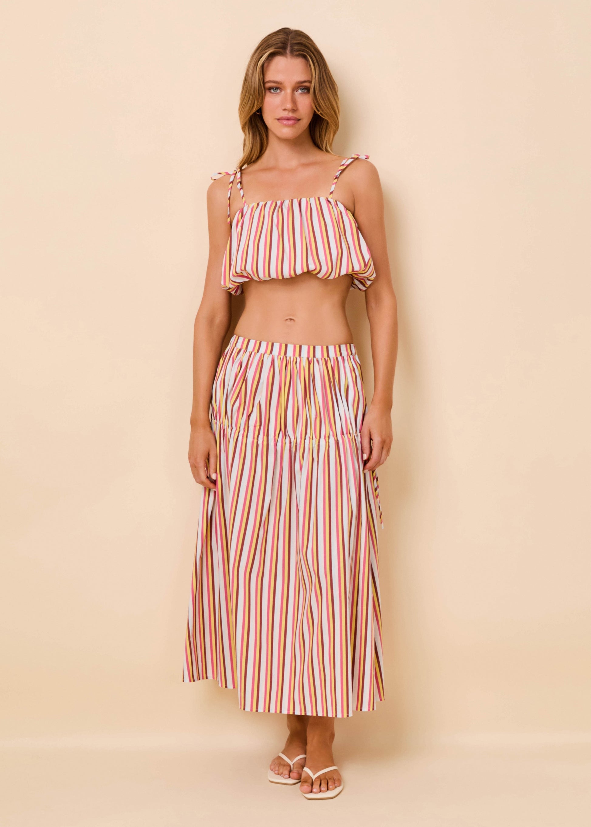 A woman modeling a two-piece set featuring a ruffled crop top and a matching maxi skirt, both with vertical pink, orange, and white stripes, set against a solid beige background.