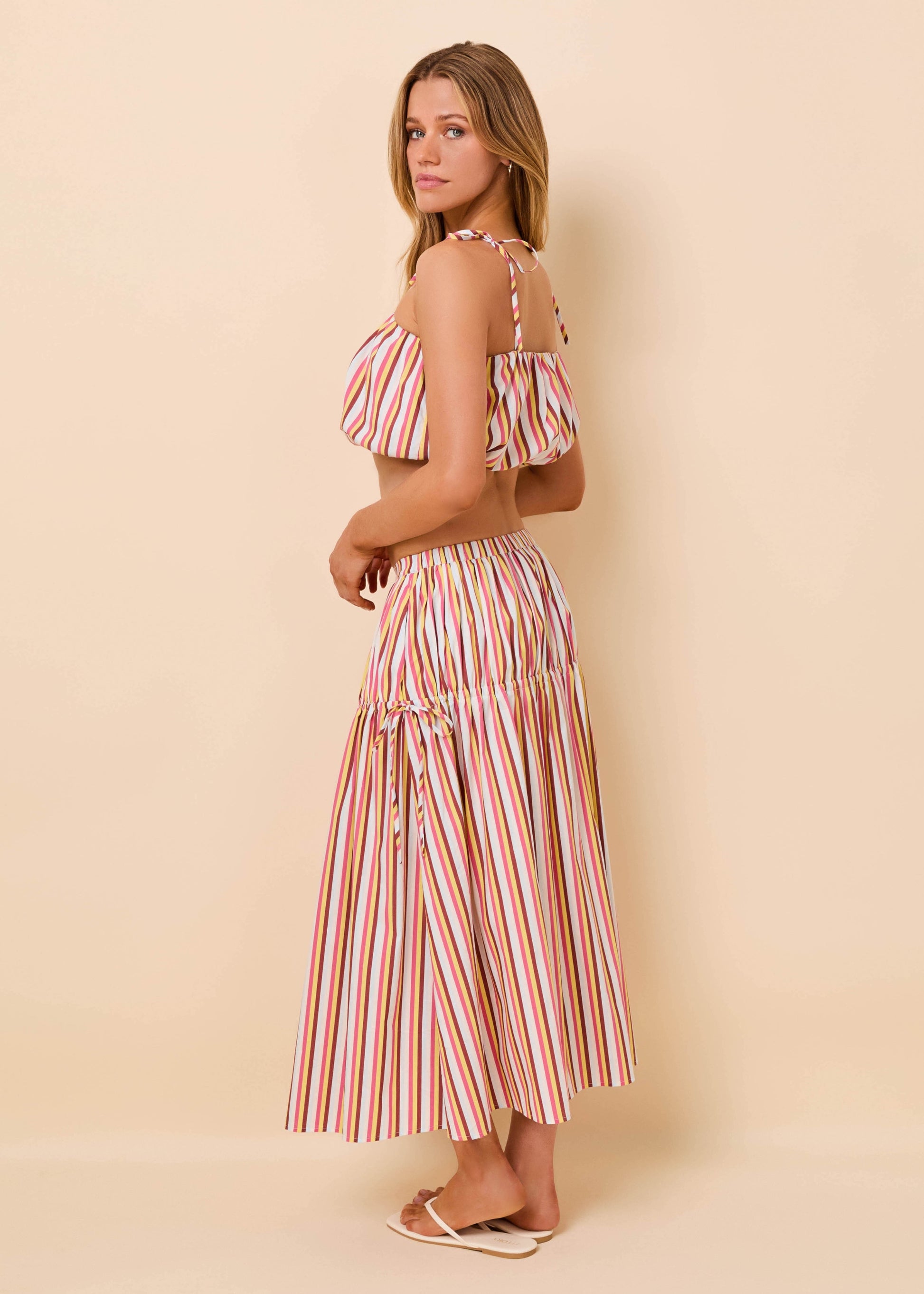 A woman modeling a two-piece striped set consisting of a cold-shoulder crop top and a matching tiered maxi skirt, set against a solid beige studio background.
