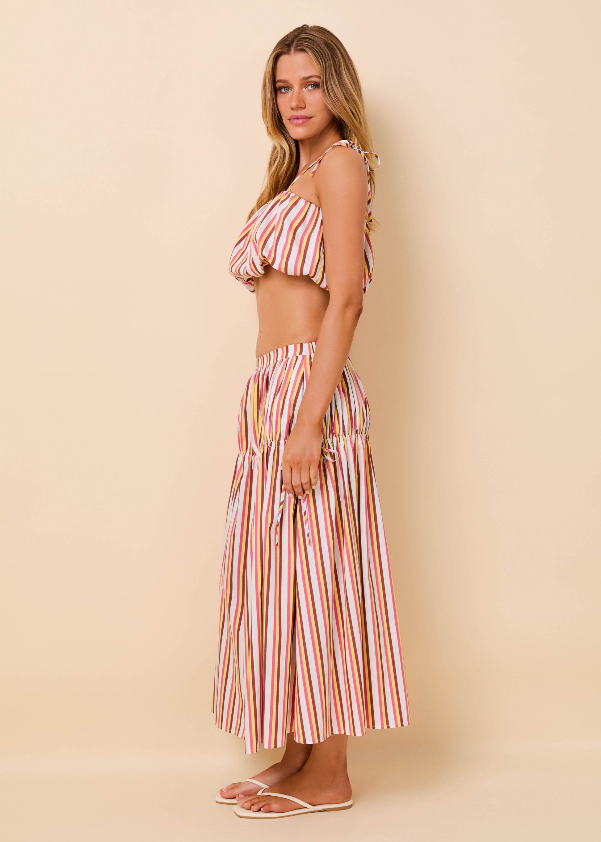 A woman modeling a two-piece striped set consisting of a halter crop top and a matching pleated maxi skirt, in pink, orange, and white vertical stripes, against a solid beige backdrop.