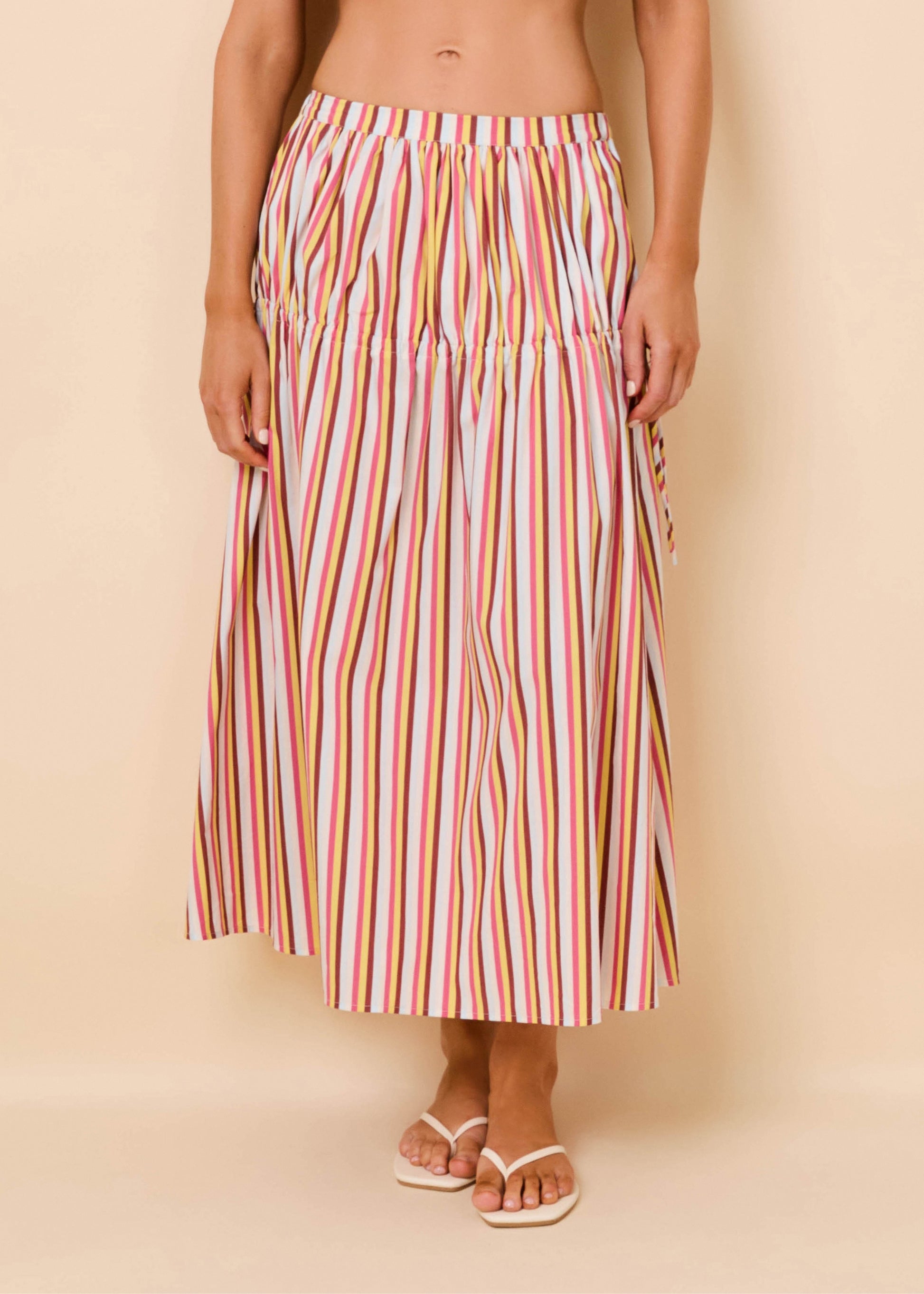 A model wearing a high-waisted, midi-length maxi skirt with vertical multicolor stripes in pink, orange, yellow, green, and blue on a white base, paired with white slide sandals, set against a solid beige studio background.
