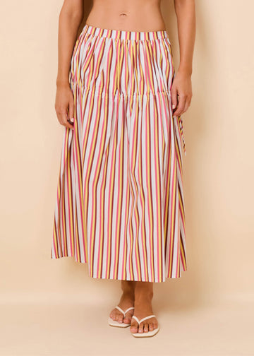 A model wearing a high-waisted, midi-length maxi skirt with vertical multicolor stripes in pink, orange, yellow, green, and blue on a white base, paired with white slide sandals, set against a solid beige studio background.