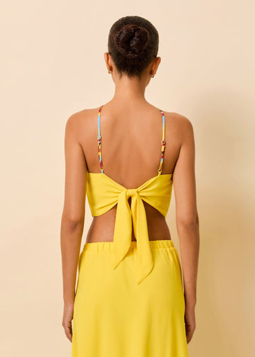 A back view of a woman modeling a bright yellow two-piece outfit consisting of a scarf top and a matching skirt with a decorative multicolor straps, posed against a plain beige wall.