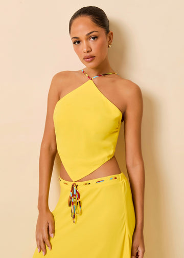 A woman modeling a bright yellow two-piece outfit consisting of a halter-style cutout top and a matching skirt with a decorative multicolor tie detail at the waist, posed against a plain beige wall.