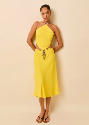 A woman wearing a bright yellow midi skirt with a tie waist and charm detail, paired with matching scarf top and black flip-flop sandals, against a solid beige studio background.