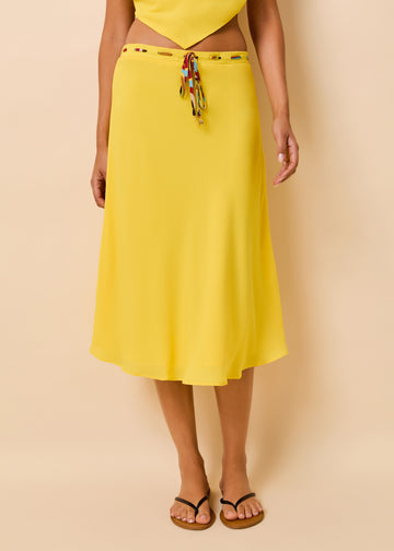A woman wearing a bright yellow midi skirt with a tie waist and charm detail, paired with black flip-flop sandals, against a solid beige studio background.
