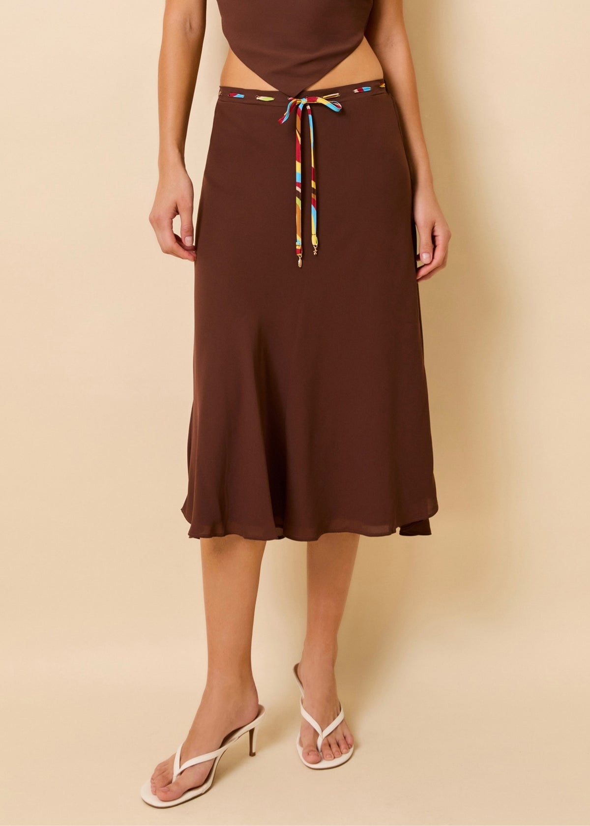 A model wearing a brown midi skirt with a colorful patterned drawstring, paired with white heeled thong sandals, against a solid beige background.