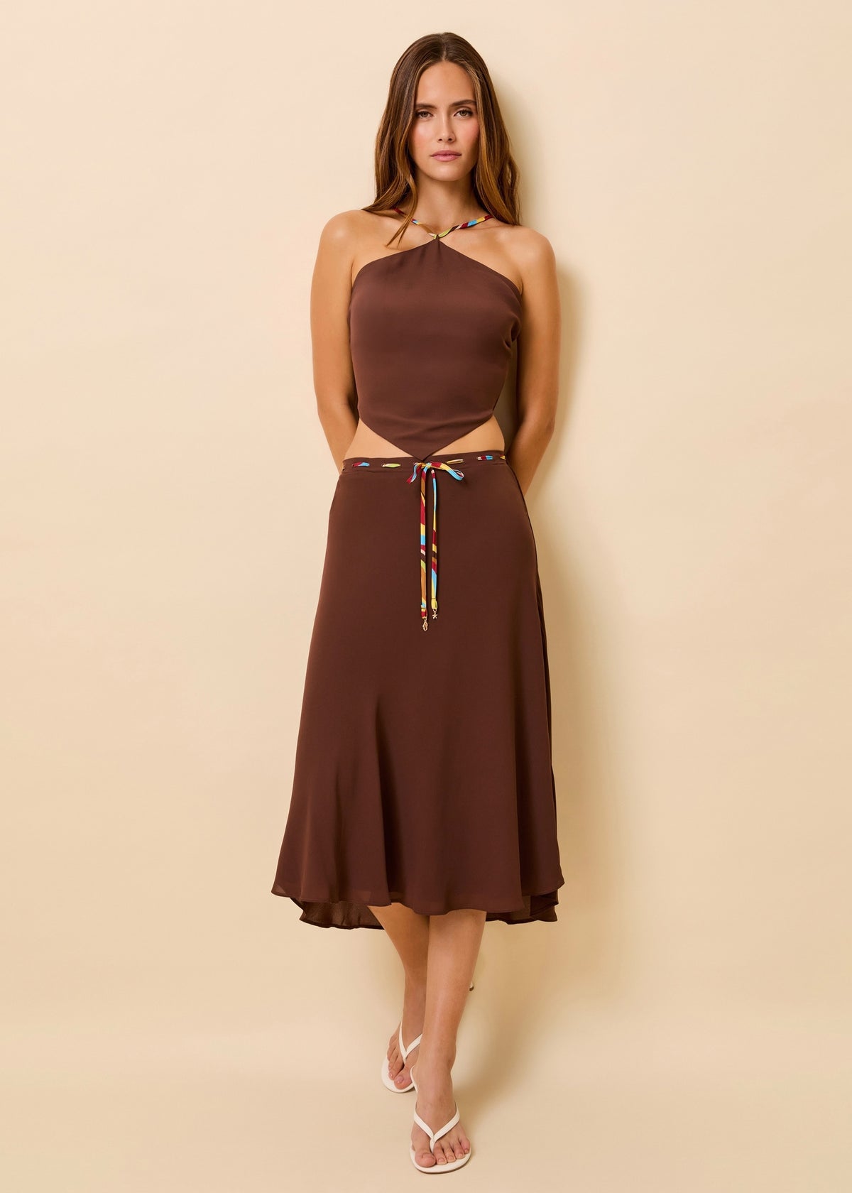 A woman modeling a brown two-piece scarf top and midi skirt set with a colorful tie detail, standing against a solid beige background.
