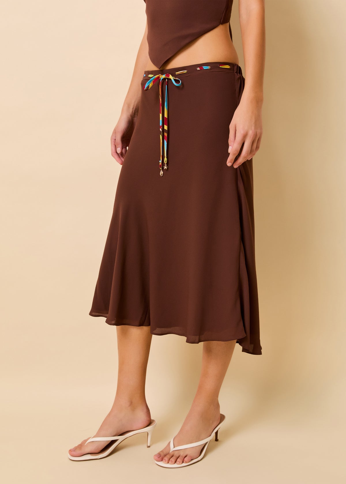 A model wearing a brown midi skirt with a colorful tie waist detail, paired with white heeled thong sandals, against a solid beige background.