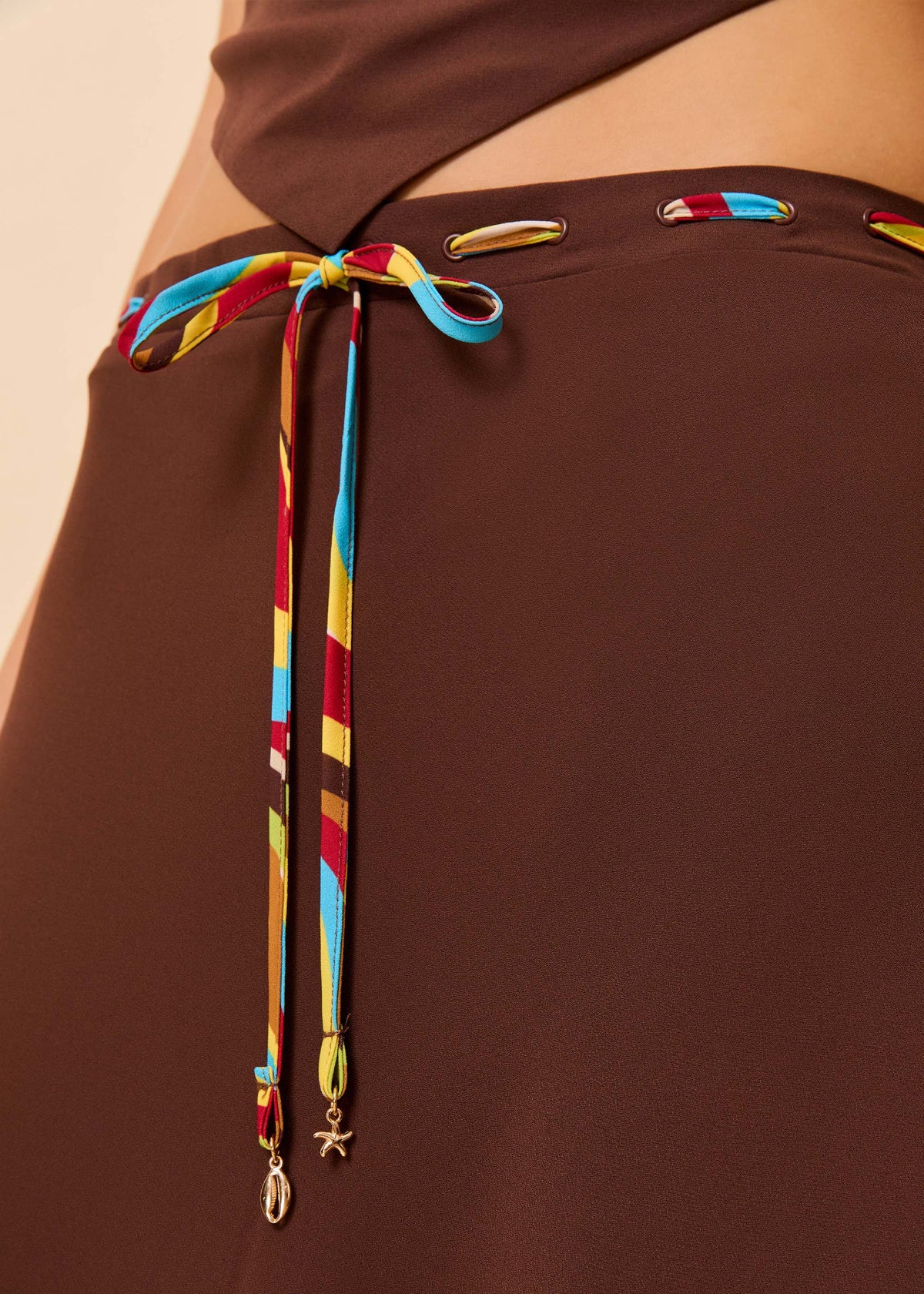 Close-up of a brown skirt with a colorful striped drawstring tied at the waist and gold-tone charms, shown on a model.