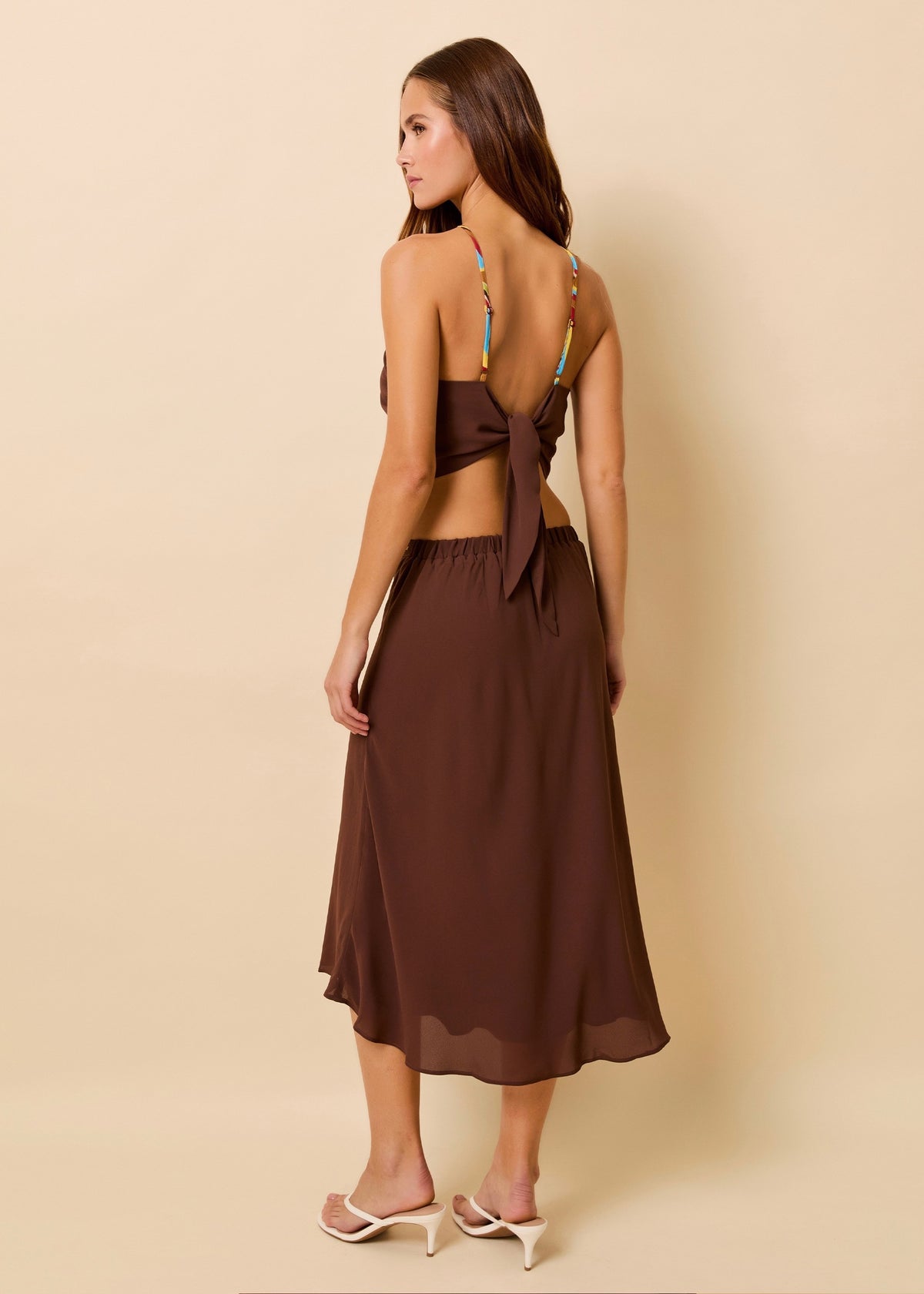 A woman modeling a brown two-piece set consisting of a scarf top with a tie-back detail and a matching midi skirt, shown from the back against a solid beige background.