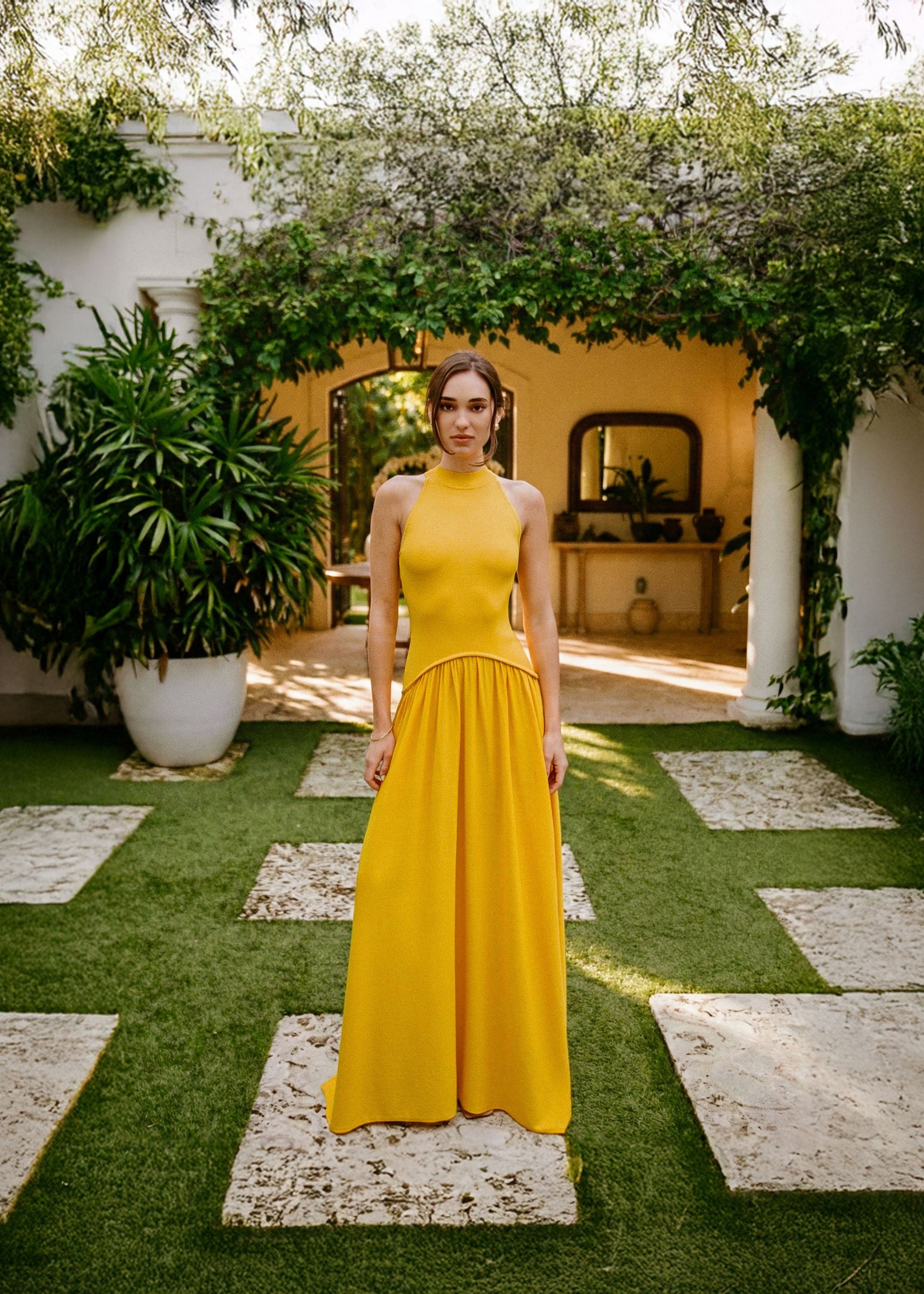 Woman in a yellow dress standing in a garden with greenery and a white building in the background.
