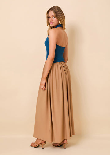 A woman modeling a sleeveless maxi dress with a fitted teal blue top and a flowing tan skirt, shown from the back against a solid beige studio background.