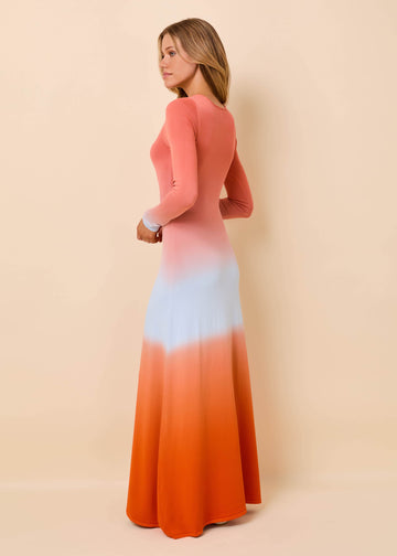 A woman modeling a long-sleeve maxi dress with a vertical ombre gradient transitioning from coral pink at the top, through pale blue in the middle, to bright orange at the bottom, set against a solid beige studio background.