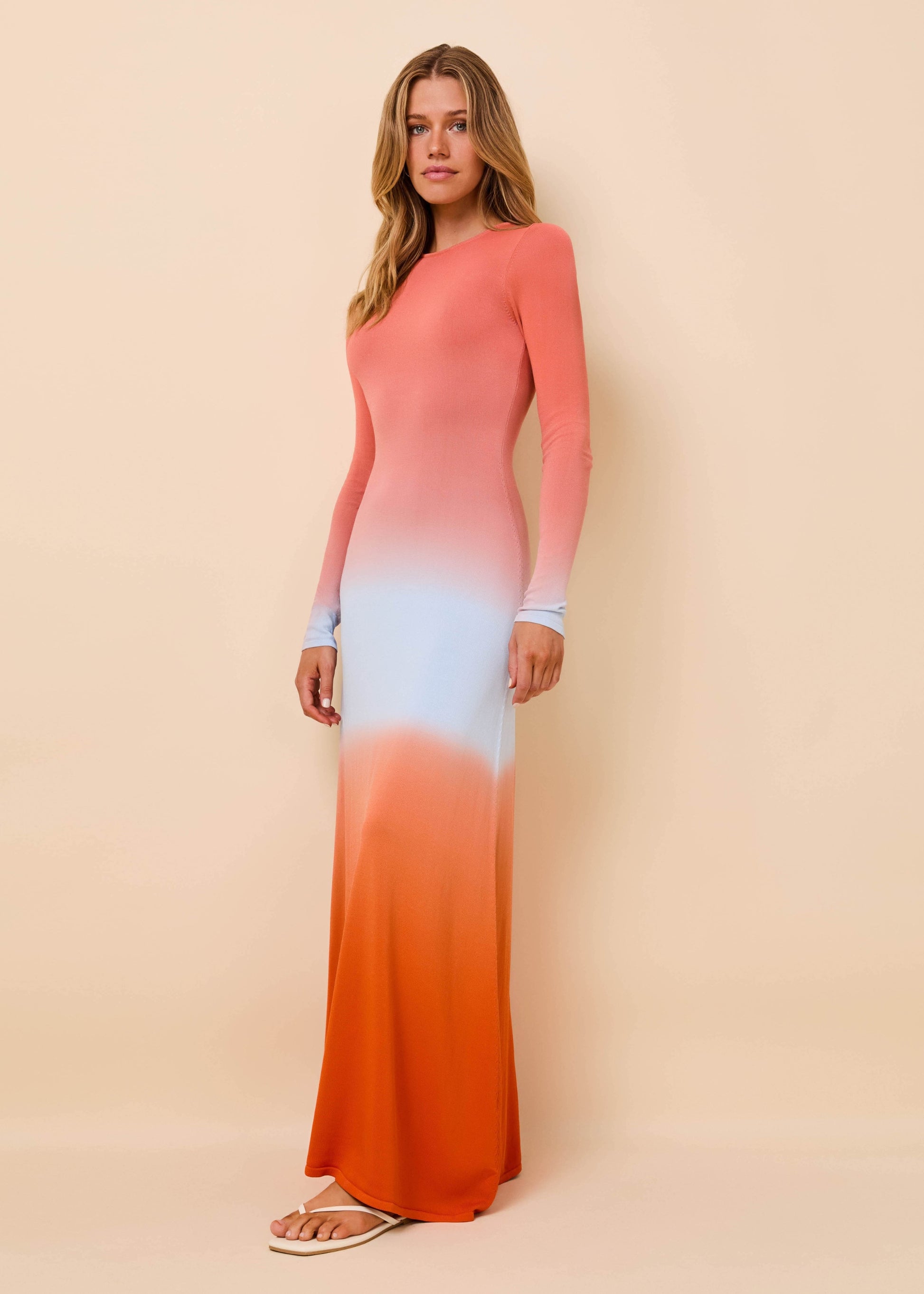A woman modeling a long-sleeve maxi dress with a vertical ombré color-block pattern in coral pink, light blue, and orange, standing against a solid beige studio background.