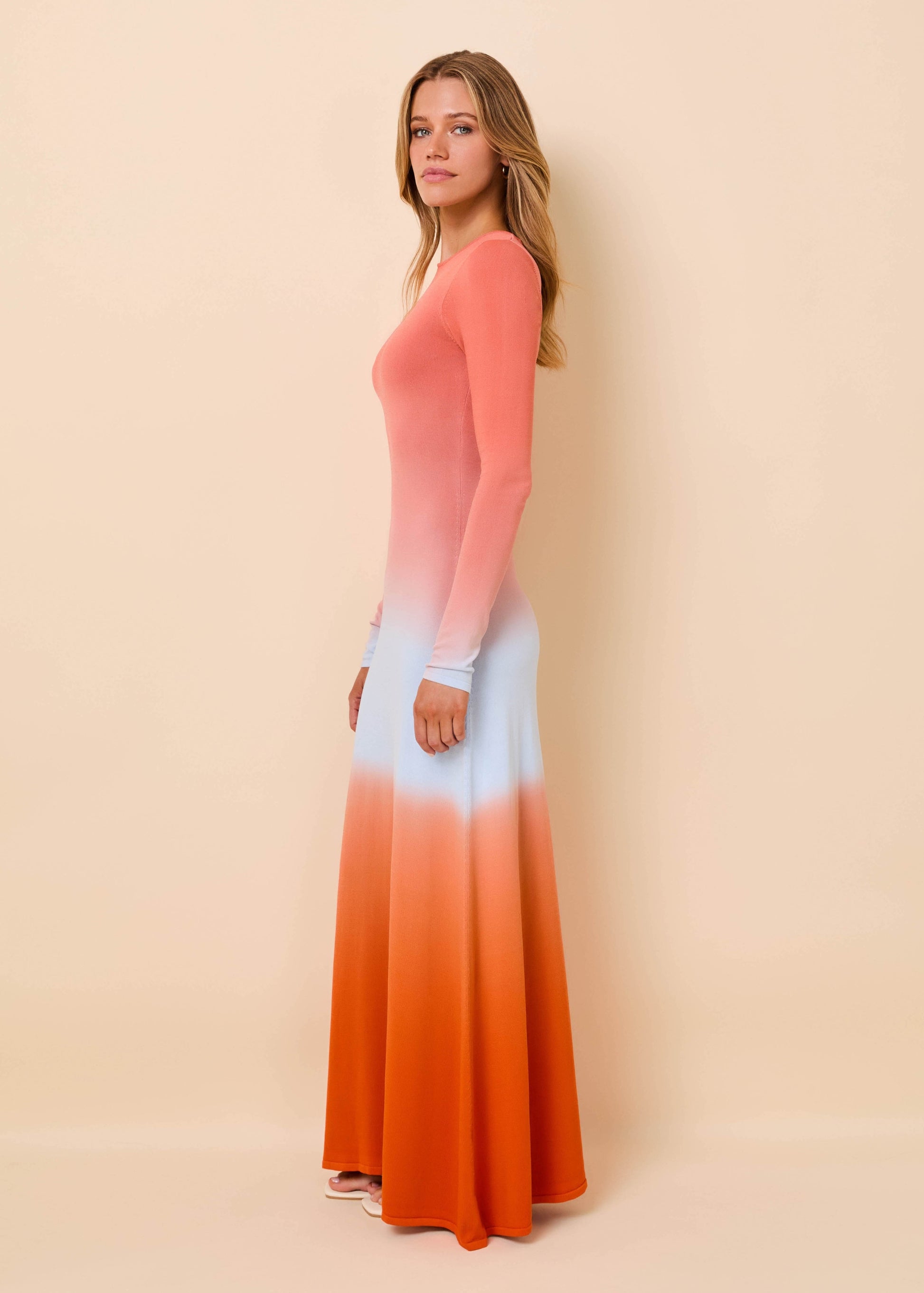 A woman modeling a long-sleeve maxi dress with a vertical ombré color-block design transitioning from coral pink at the top, through white in the middle, to bright orange at the hem, set against a solid beige studio background.