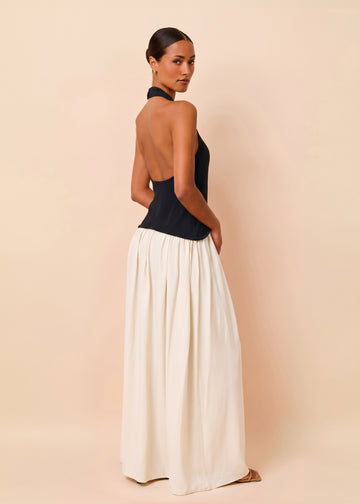 A woman modeling a sleeveless halter-neck maxi dress with a dark navy blue bodice and a flowing off-white skirt, shown from a rear three-quarter view against a solid beige studio backdrop.