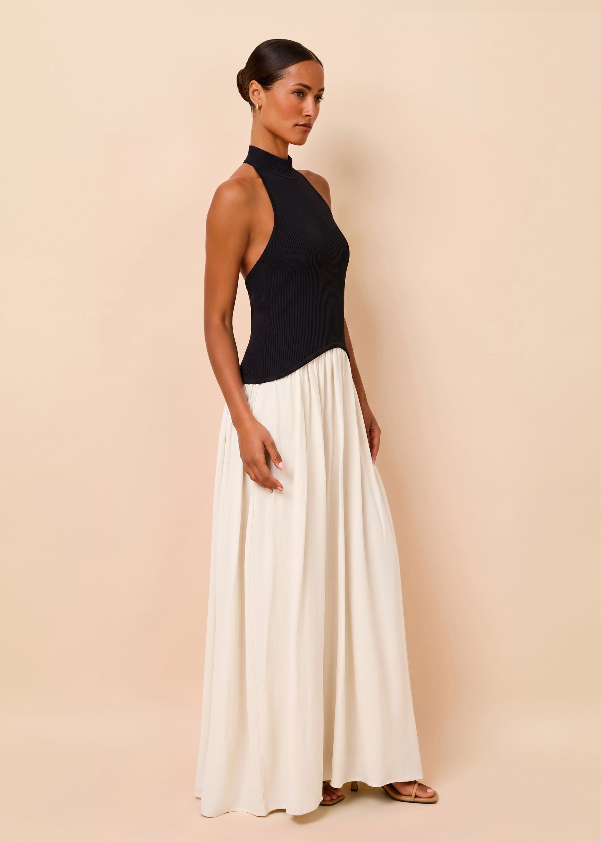 A woman modeling a sleeveless black mock-neck top paired with a long, flowing white skirt, standing against a solid beige studio backdrop.