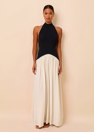 A woman modeling a sleeveless maxi dress with a black halter-style bodice and a flowing cream skirt, standing against a plain beige studio background.