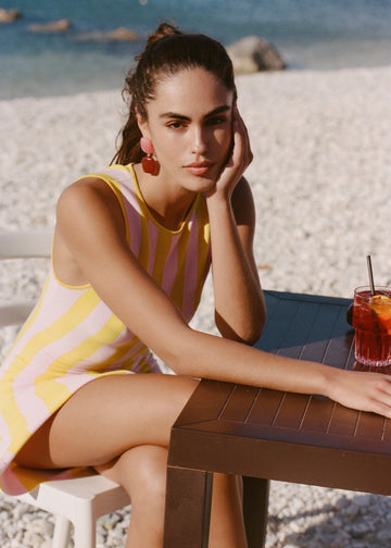A woman sitting at a dark wooden table on a beach, wearing a yellow and pink striped sleeveless mini dress. A glass of iced tea with a straw is on the table next to her.