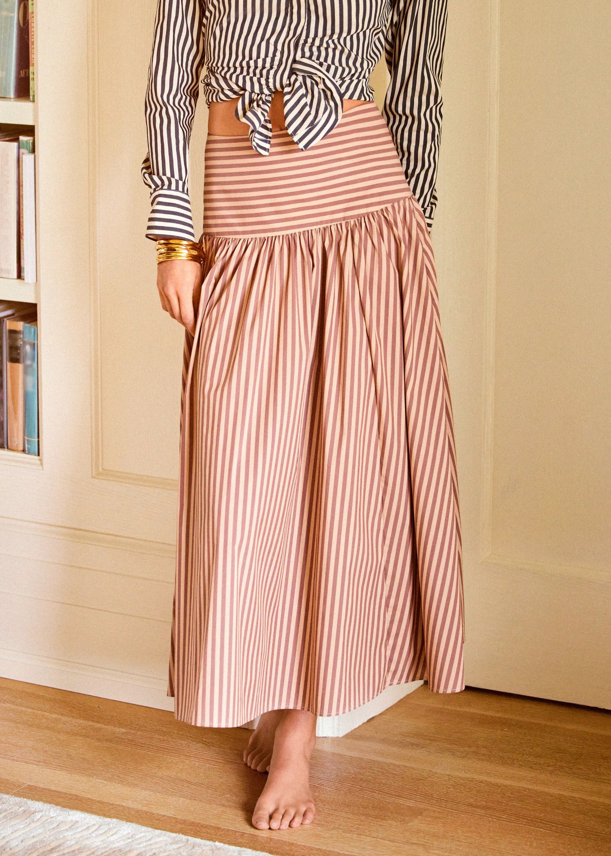 A woman wearing a high-waisted, long maxi skirt with vertical pink and white stripes, paired with a matching striped tie-front top. She is standing barefoot on a wooden floor in a warm, neutral-toned room.