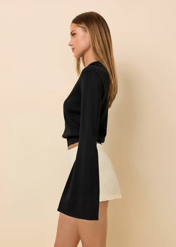 A woman shown in side profile wearing a black long-sleeve crop top with a flared bell sleeve and a white skirt, standing against a plain beige studio background.