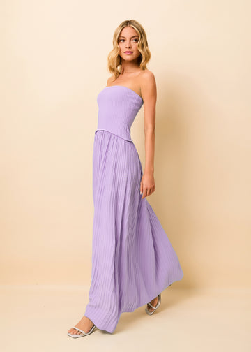 A woman modeling a lavender strapless maxi dress with a ribbed bodice and flowing pleated skirt, standing against a solid beige studio backdrop.