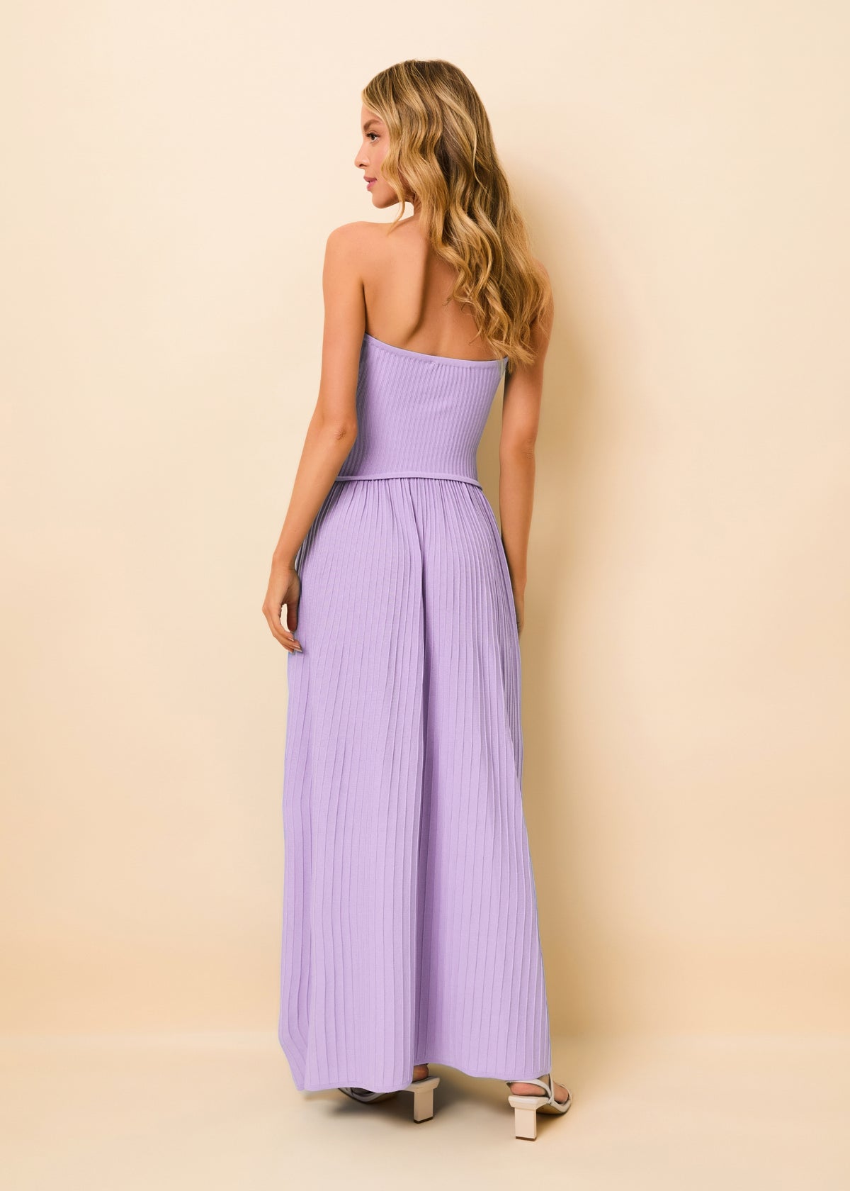 A woman shown from the back wearing a lavender ribbed strapless maxi dress with a structured bodice and flowing skirt, standing against a solid beige studio background.