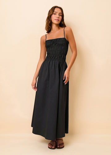 A woman modeling a black midi dress with spaghetti straps, a smocked bodice, and a flared skirt, standing against a solid beige background.