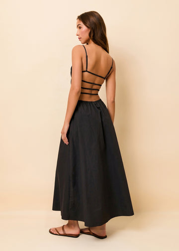 A woman modeling a black maxi dress from the back, featuring a strappy open-back bodice and a full gathered skirt, set against a solid beige studio background.